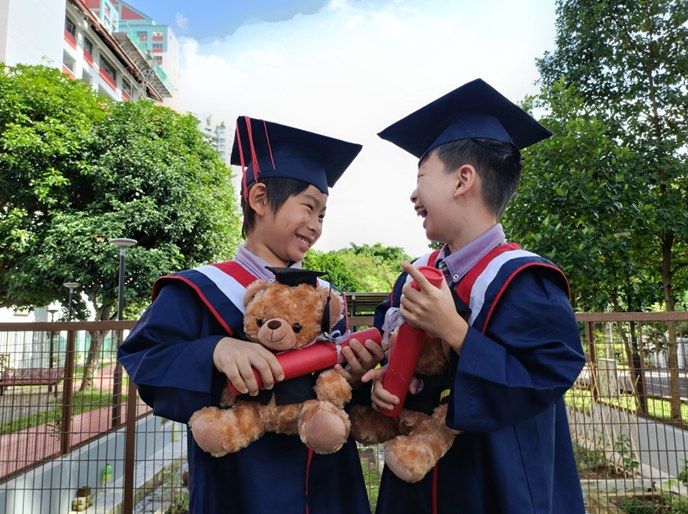 Two graduating boys in caps and gowns laughing together while holding their diplomas and teddy bears
