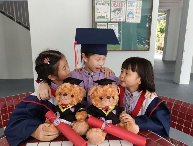 Three graduating children in caps and gowns hugging and smiling together with their teddy bears